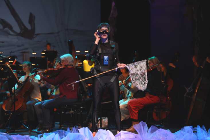 Eine Schauspielerin in einem Taucheranzug steht auf der Bühne. Sie hält einen Käscher in der Hand und trägt eine Taucherbrille. Hinter ihr sitzt das Orchester.