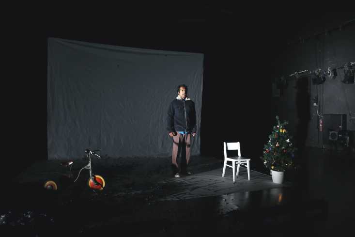 Die Bühne ist dunkel und karg. Links steht ein Dreirad, rechts ein weißer Stuhl und ein kleiner Weihnachtsbaum. In der Mitte steht mit hängenden Armen und in Winterjacke der Schauspieler.