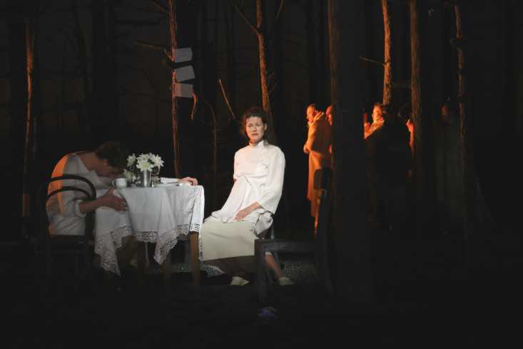 Eine schwangere Frau in einem weißen Kleid sitzt an einem weiß gedeckten Tisch. Über den Tisch gebeugt sitzt ein Mann, sein Blick geht auf den Boden. Im Dunkel hinter ihnen stehen mehrere Personen.