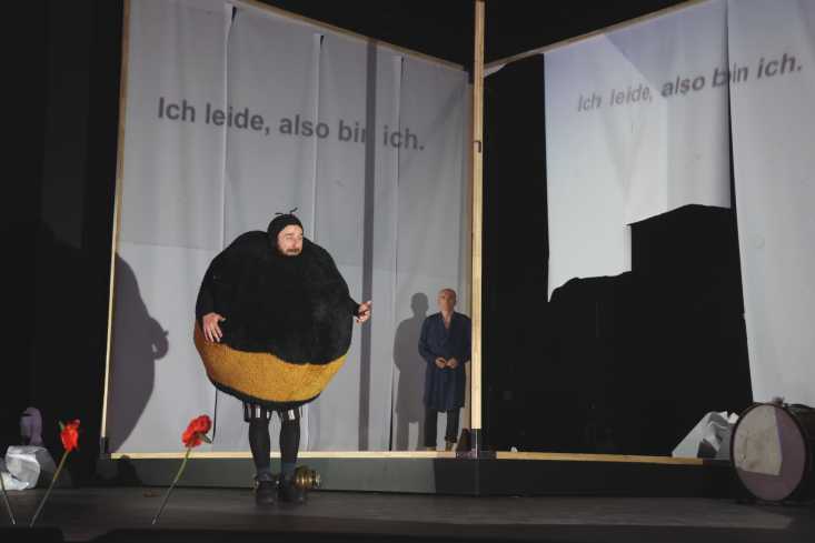 Ein Schauspieler in einem plüschigen Bienenkostüm steht auf der Bühne. Auf eine Papierbahn hinter ihm ist ein Satz projeziert: Ich leide, also bin ich. 