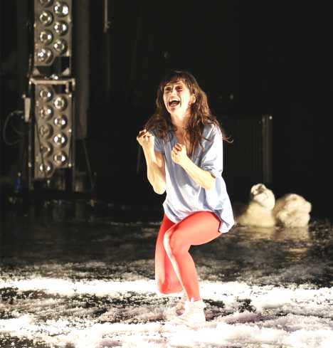 Eine Frau steht mit gebeugten Knien und geballten Fäusten auf der Bühne. Sie schreit. Um sie herum liegt Kunstschnee auf dem Boden.
