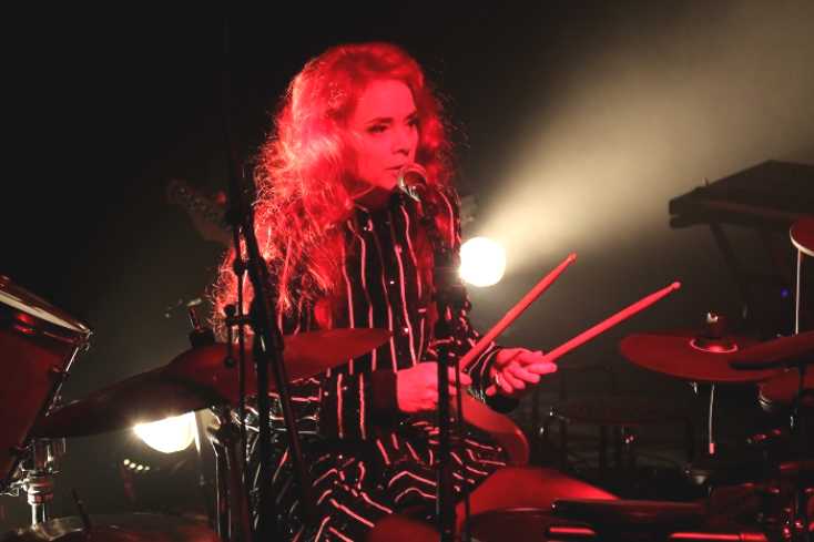 Eine Frau sitzt am Schlagzeug. Sie singt und hält die die Sticks in den Händen.