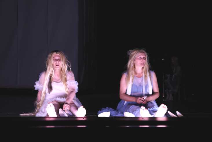 Zwei Schauspielerinnen sitzen auf dem Boden. Um sie herum ist alles dunkel. Beide tragen Kleider und blonde Langhaarperücken. 