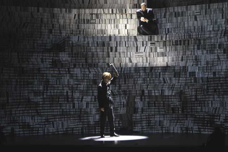 Oben im Bühnenbild sitzt ein Sänger in einem schwarzen Gewand. Die Bühne ist dunkel, er schaut zu einem anderen Sänger, der unten in einem Lichtkreis steht, herunter. 