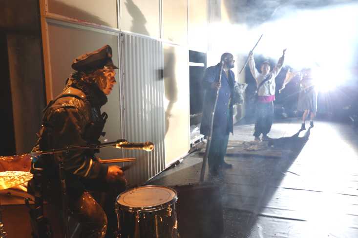 Im Vordergrund steht eine Frau in Uniform an einem Schlagzeug. Hinten stehen drei Personen, einer hält die Arme hoch, eine andere schwenkt mit einer Art Fahne.