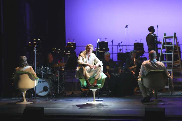 Drei Menschen sitzen in Drehstühlen auf der Bühne, die beiden äußeren Personen drehen dem Publikum den Rücken zu. Der Sänger in der Mitte schaut stumm zu uns. Im lila beleuchteten Hintergrund sitzt die Band.
