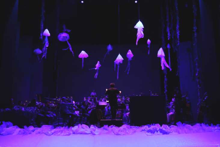 Die Bühne ist dunkel. Im Schwarzlicht hängen leuchtende Quallen aus dem Bühnenhimmel. Darunter sitzt das Orchester.