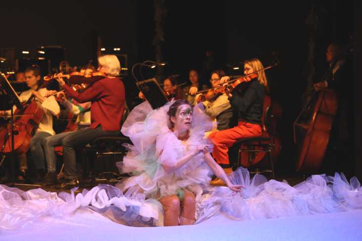 Eine Sängerin ganz in rosafarbenen Tüll gehüllt sitzt vorn auf der Bühne. Hinter ihr sitzt das Orchester. 