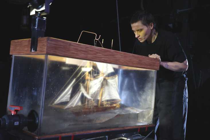 Eine Schauspielerin schaut in einen Glaskasten, in dem ein großes Segelschiff schwimmt.