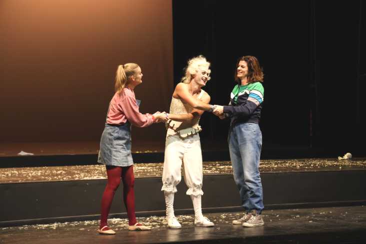 Drei Menschen stehen auf der Bühne. Der Mann in der Mitte trägt ein barockes Kostüm mit Lockenperücke. Zwei Sängerinnen halten ihn lachend fest.