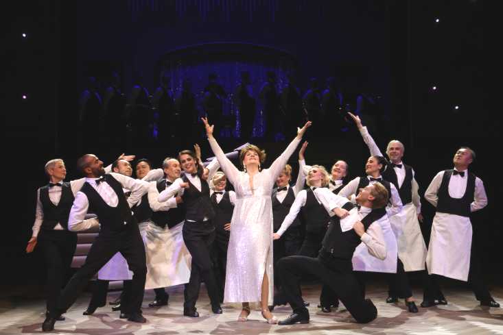 Eine Frau in einem leuchtend weißen Kleid steht mit erhobenen Armen auf der Bühne. Um sie herum steht und tanzt ein Chor aus Kellnern. Alle schauen zu ihr.