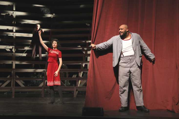 Hinten auf der Bühne steht eine Frau in einem roten Kleid. Vorn steht ein Mann hinter dem Bühnenvorhang, so dass sie ihn nicht sehen kann.