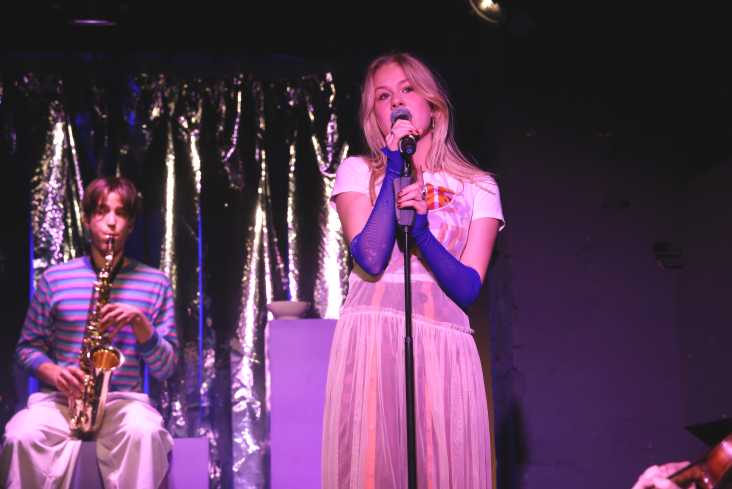 Ein junger Akteur spielt Saxofon, eine junge Akteurin steht an einem Mikrofonständer und singt.