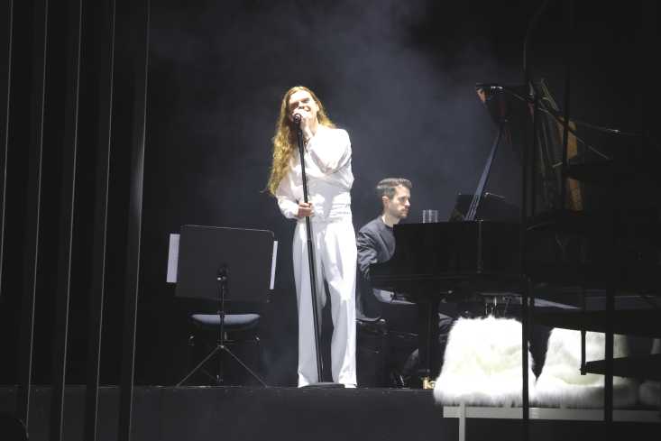 Ein Sänger steht auf der Bühne und trägt einen weißen Anzug. Im Hintergrund sitzt ein Pianist.