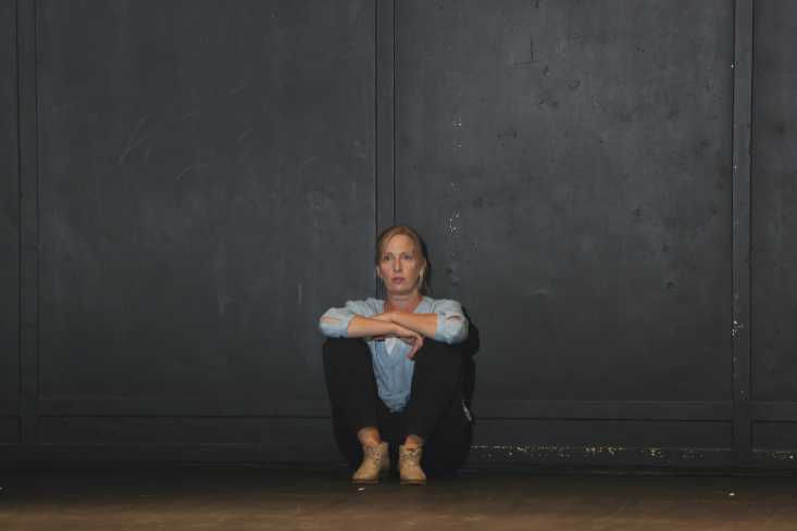 Die Schauspielerin sitzt allein an eine schwarze Wand gelehnt mit angezogenen Beinen auf dem Boden.