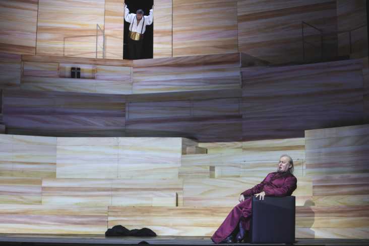 Gesamtaufnahme der Bühne. Auf einem Sessel unten rechts sitzt ein Säner in einem langen, purpurfarbenen Gewand. Oben in der Bühnenmitte steht ein Sänger in einer geöffnetem Tür und schaut zu ihm runter. 