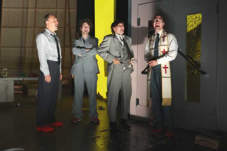 Vier Schauspieler stehen nebeneinander und lachen mit geöffneten Mündern. Drei tragen einen Anzug, einer ist gekleidet wie ein Priester.