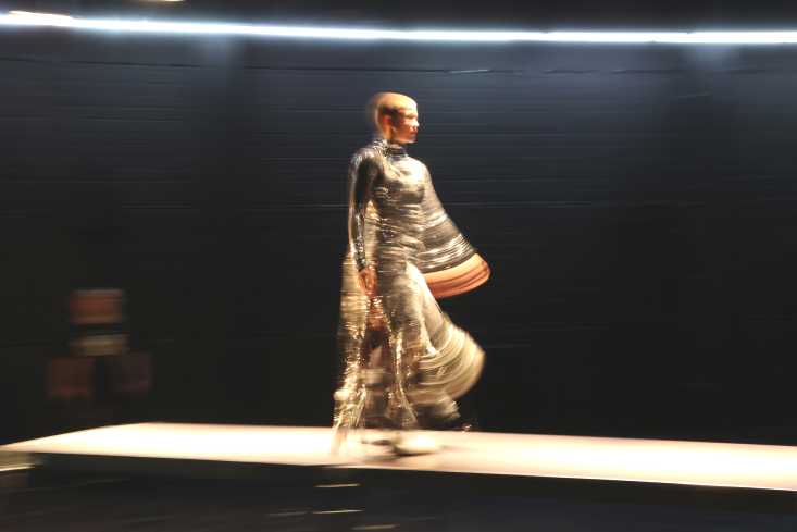 Eine Frau in einem metallisch-glänzendem Outfit geht über einen Laufsteg. Das Bild ist leicht verschwommen.