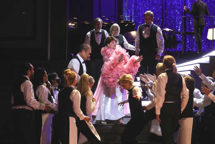 Eine Frau in einem weißen Kleid mit einer großen Federboa schreitet eine Showtreppe runter. Sie wird dort von vielen Kellnern empfangen.