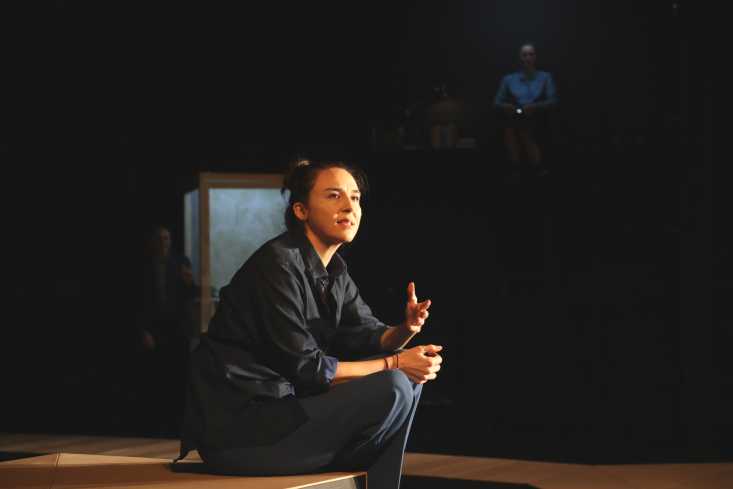 Eine Frau sitzt auf einer Treppenkante und gestikuliert. Die Bühne hinter ihr liegt im Dunkeln.