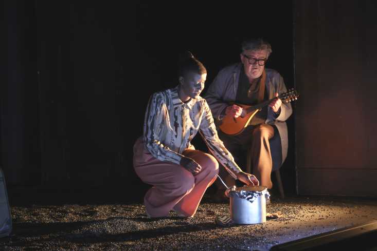 Eine Frau hockt vor einem großen Topf. Hinter ihr sitzt ein Mann und spielt Mandoline.