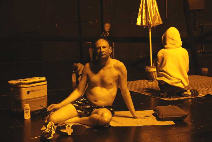Ein Mann sitzt mit einer Badehose bekleidet auf einem Handtuch. Hinter ihm sind zwei lebensgroße Puppen zu sehen, die picknicken.