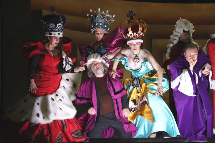 Ein Mann kniet auf der Bühne. Er ist umgeben von drei Frauen in opulenten Königinnenkostümen.