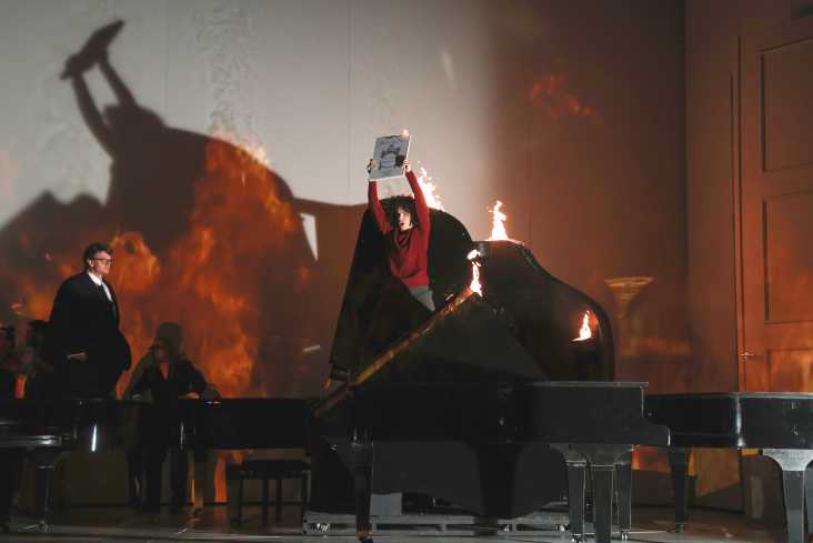 Auf der Bühne stehen mehrere Flügel, zwei sind aufgeklappt. In einem steht eine Person mit einem brennenden Manuskript. Auch die Flügel brennen. Auf die Wand hinten sind Flammen projiziert.