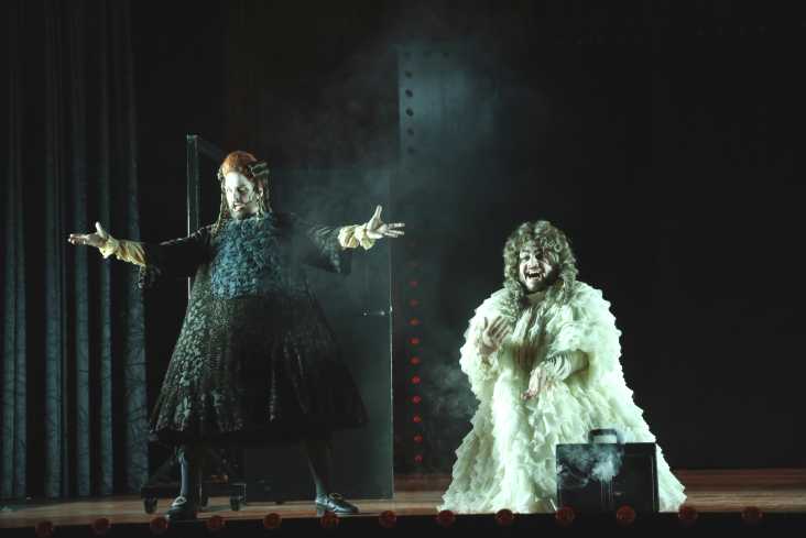 Ein dunkel gekleideter Sänger steht mit weit ausgebreiteten Armen auf der Bühne. Neben ihm kniet ein anderer Sänger mit einer Aktentasche. Er trägt einen weißen Mantel.