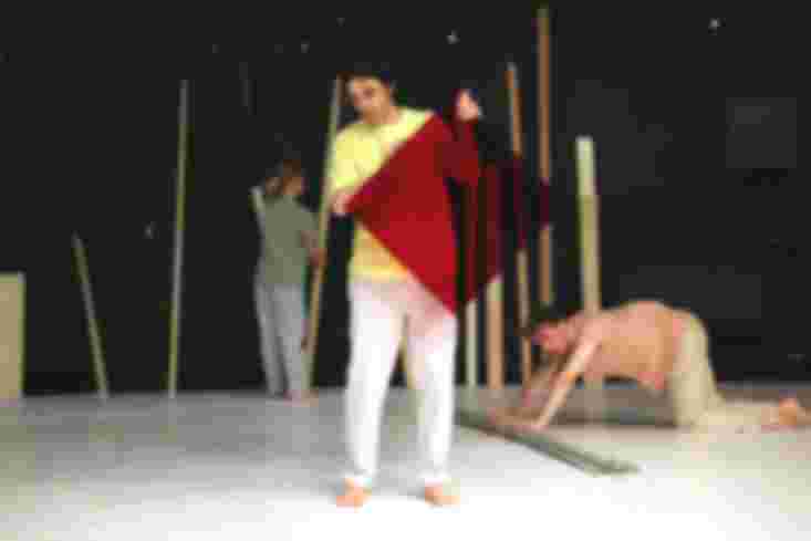 Ein Tänzer steht vorn. Er hält eine transparente, rote Scheibe in den Händen. Hinter ihm schiebt ein anderer Tänzer dünne Rohre über den Boden. Eine Tänzerin nimmt eine Holzlatte.