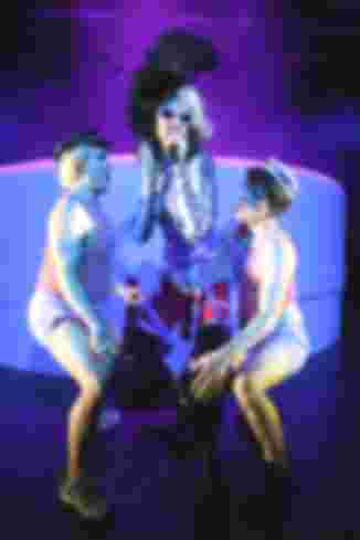Eine Person mit Sonnenbrille und schwarzem Federhut singt. Zwei andere Personen tanzen sie von beiden Seiten an.