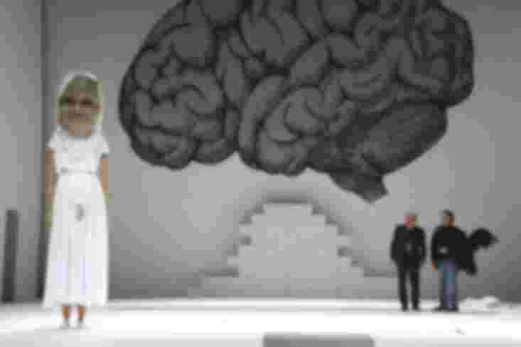 Die Comiczeichnung eines Gehirns ist an der Rückwand zu sehen. Vorn steht eine sehr große Puppe, sie trägt ein weißes Kleid. Hinten stehen zwei Männer, einer von ihnen trägt ein Vogelkostüm.