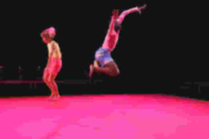 Ein Tänzer schwebt etwa einen Meter über dem Boden mitten im Sprung, seine Beine sind angezogen, seine Arme nach oben gestreckt. Eine Tänzerin hinter ihm macht kleine Sprünge auf der Stelle. Ihre Haare fliegen.