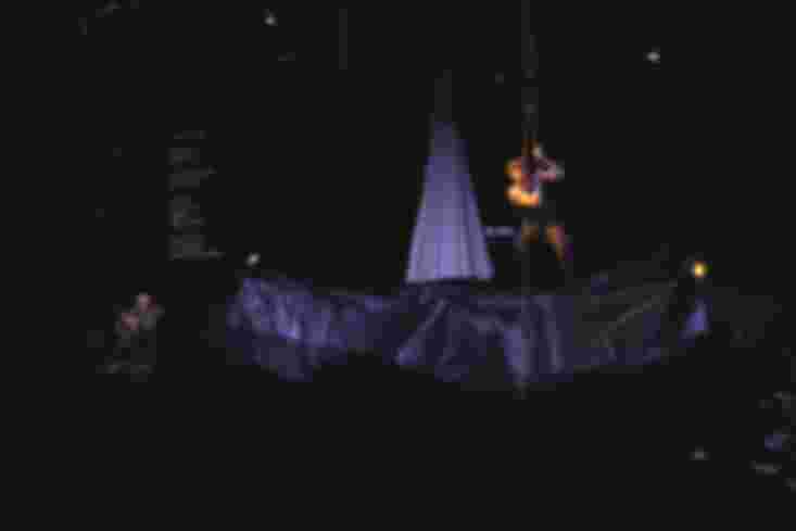 Die Bühne ist dunkel und voller schwarzem Papier. Hinten spielt ein Mann Gitarre, ein anderer Mann sitzt auf einer Art Schaukel in der Luft.