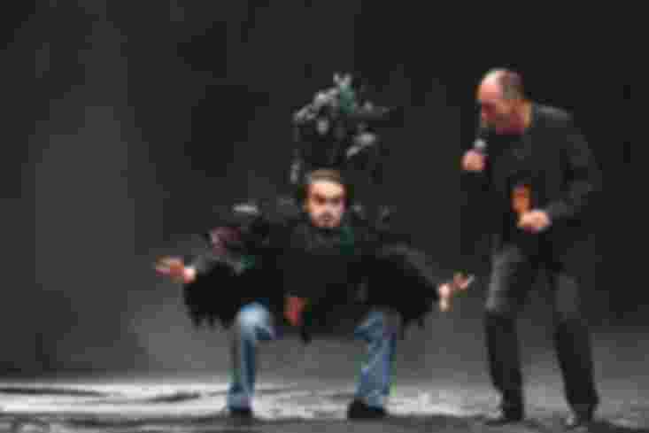 Ein Mann im Vogelkostüm steht in der Hocke mit gespreizten Armen auf der Bühne. Neben ihm steht ein zweiter Mann.