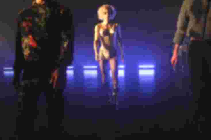 Drei Menschen kommen auf das Publikum zu. In der Mitte ein Schauspieler mit blonder Madonna-Perrücke und hochhackigen Stiefeln. Er trägt ein Mieder und Strumpfbänder. Er ist als einziger ganz im Bild und auch im Scheinwerferlicht.