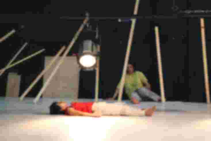 Eine Tänzerin liegt auf dem Rücken auf dem Boden. Nah über ihr leuchtet ein Scheinwerfer. An der Wand hinten sitzt ein Tänzer.