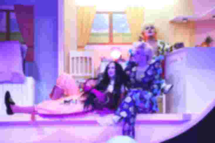 Drei Personen sitzen und liegen in einer übergroßen Puppenküche. Sie tragen schrille Kostüme und Perrücken.