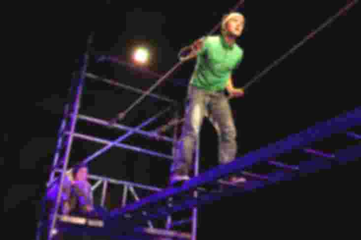 Ein Schauspieler läuft oben auf dem Gerüst über eine Brücke. An deren einem Ende sitzt mit angezogenen Beinen eine Schauspielerin und schaut ihm zu.