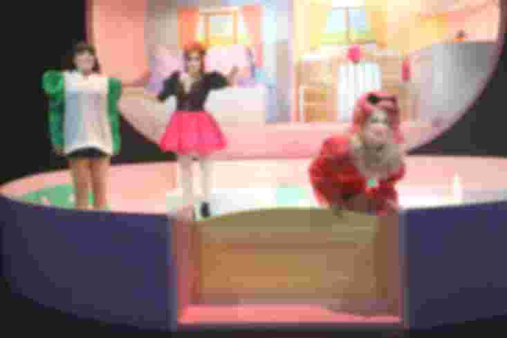 Drei Personen stehen im Garten des Bühnenbild-Puppenhauses. Sie sind stark geschminkt und teilweise grell gekleidet. 