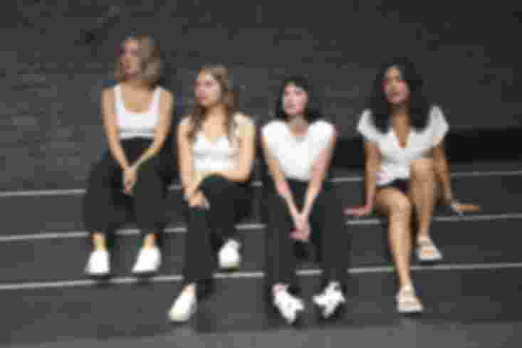 Vier junge Frauen sitzen nebeneinander auf der Treppe. Alle tragen weiße T-Shirts und schwarze Hosen.