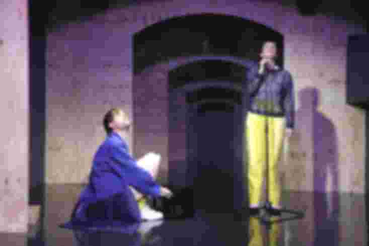 Ein Schauspieler hockt vor einem sehr kleinen Piano, eine Schauspielerin steht am Mikrofon. Beide schauen nach oben.
