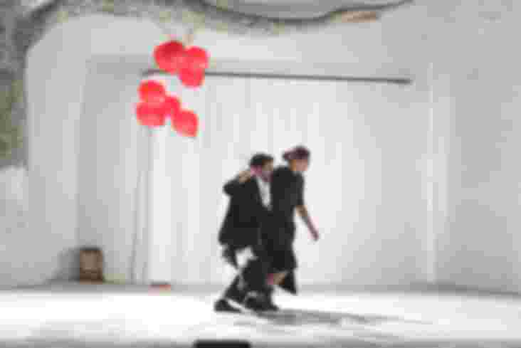 Ein Mann und eine Frau laufen über die Bühne. Der Mann hält acht rote Luftballons in der Hand.