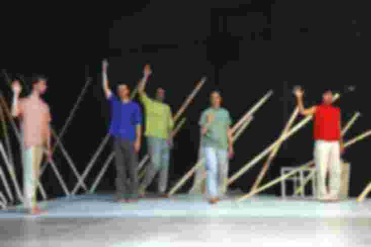 Fünf Menschen stehen auf der Bühne. Vier von ihnen haben einen Arm gehoben. Im Hintergrund lehnen Holzlatten an der Wand.