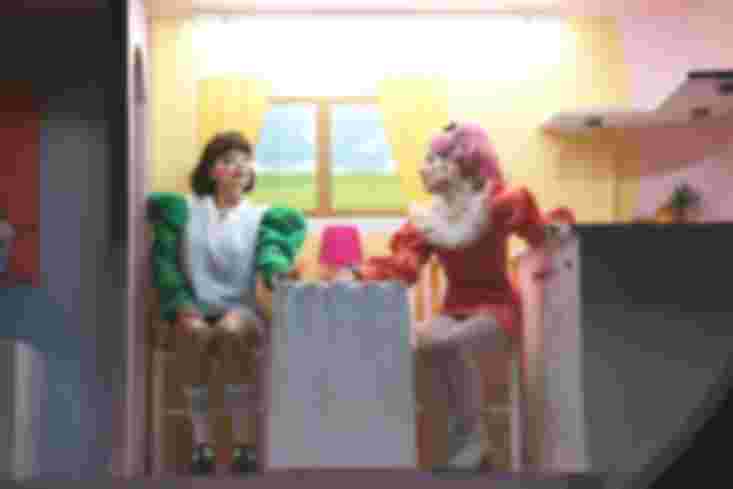 Zwei Personen sitzen in der Puppenküche auf zwei kleinen Stühlen an einem Tisch.