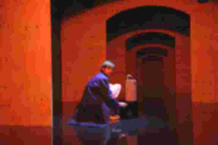 Ein Schauspieler hockt vor einem winzigen Klavier. Auf dem steht eine Trinktüte, die viel höher ist als es selbst.