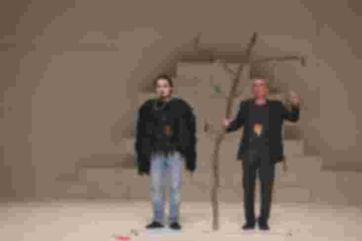 Zwei Männer stehen auf der Bühne, der eine hält einen eher dünnen Ast zwischen sie. Von dem baumeln zwei Stricke.