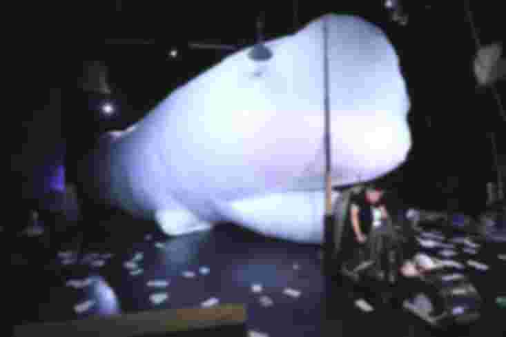 Ein riesiger weißer ausgeblasener Wal liegt auf der Bühne. Vor ihm sitzt eine Frau auf einem fahrradähnlichen Gefährt.