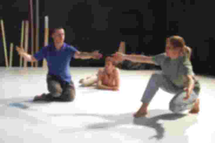 Ein Tänzer kniet auf dem Boden. Neben ihm hockt eine Tänzerin. Sie haben ihre Hände nacheinander ausgestreckt ohne sich zu berühren. Ein Tänzer liegt hinter ihnen auf dem Boden und betrachtet sie.