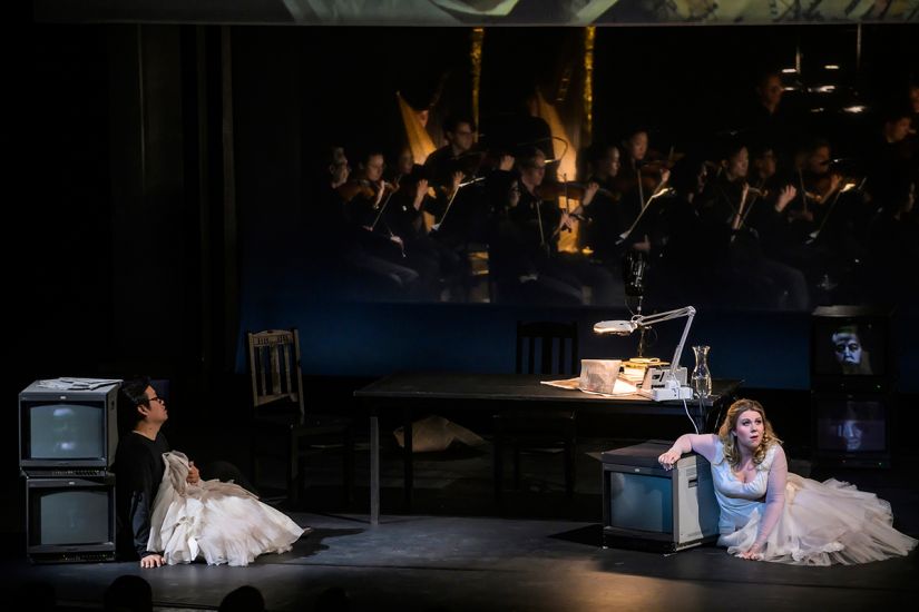 TITLE: Aida
URHEBER: Bettina Stöß Radames lehnt sich an zwei Bildschirme auf der Bühne, Aida kniet in einem Brautkleid am Boden. Hinter ihnen Radames' Schreibtisch, dahinter das Orchester der Deutschen Oper Berlin.